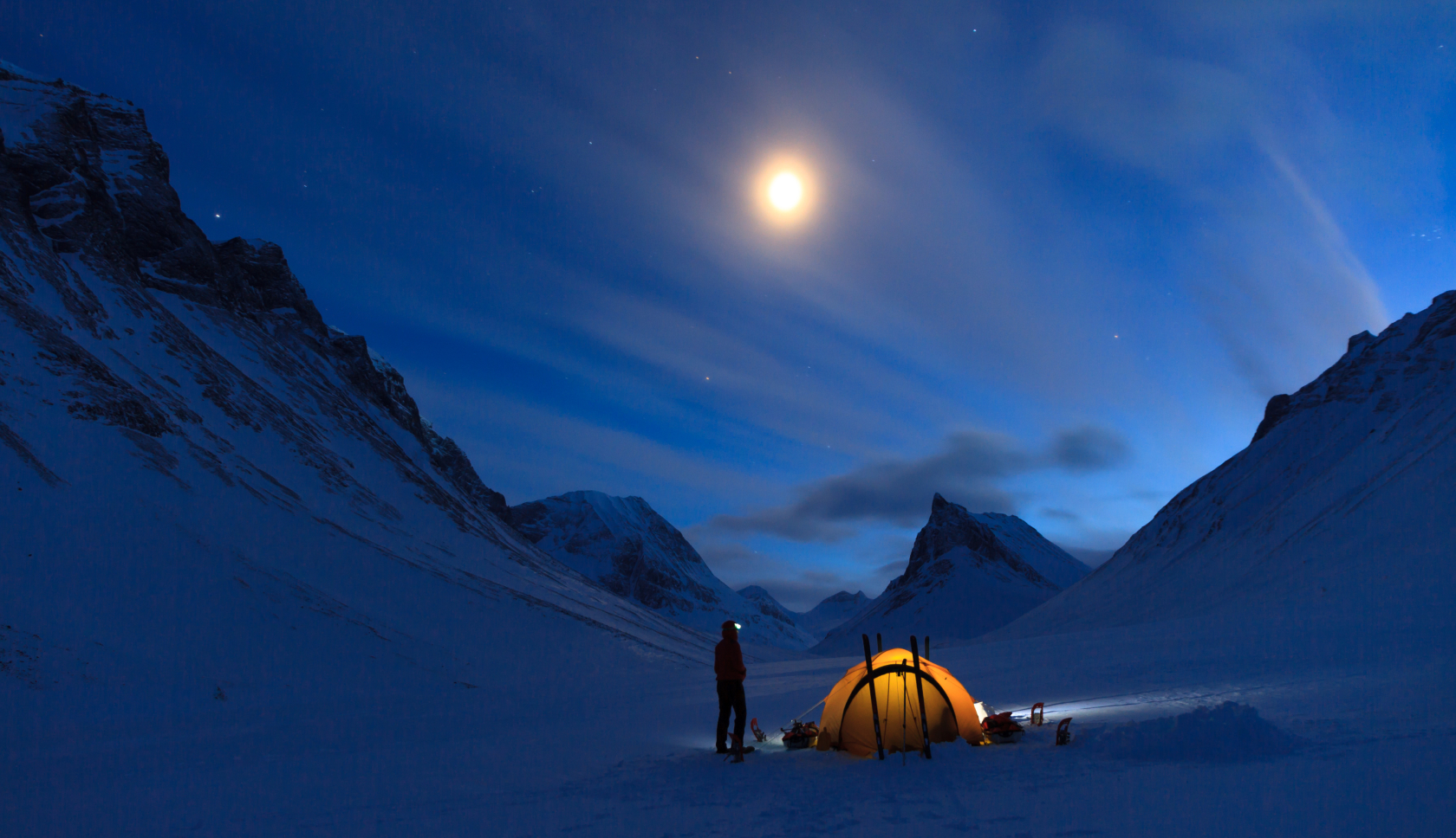 man naast tentje in besneeuwde vallei kijken naar de heldere maan in het donker