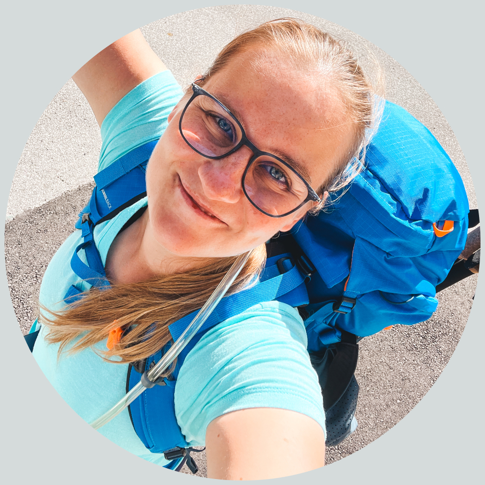 Een vrouw met een bril en een rugzak lacht blij in de camera