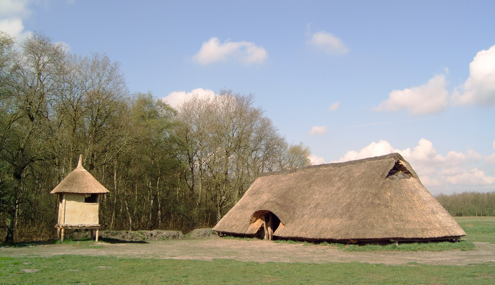Een ijzertijdboerderij op het gras bij bomen.