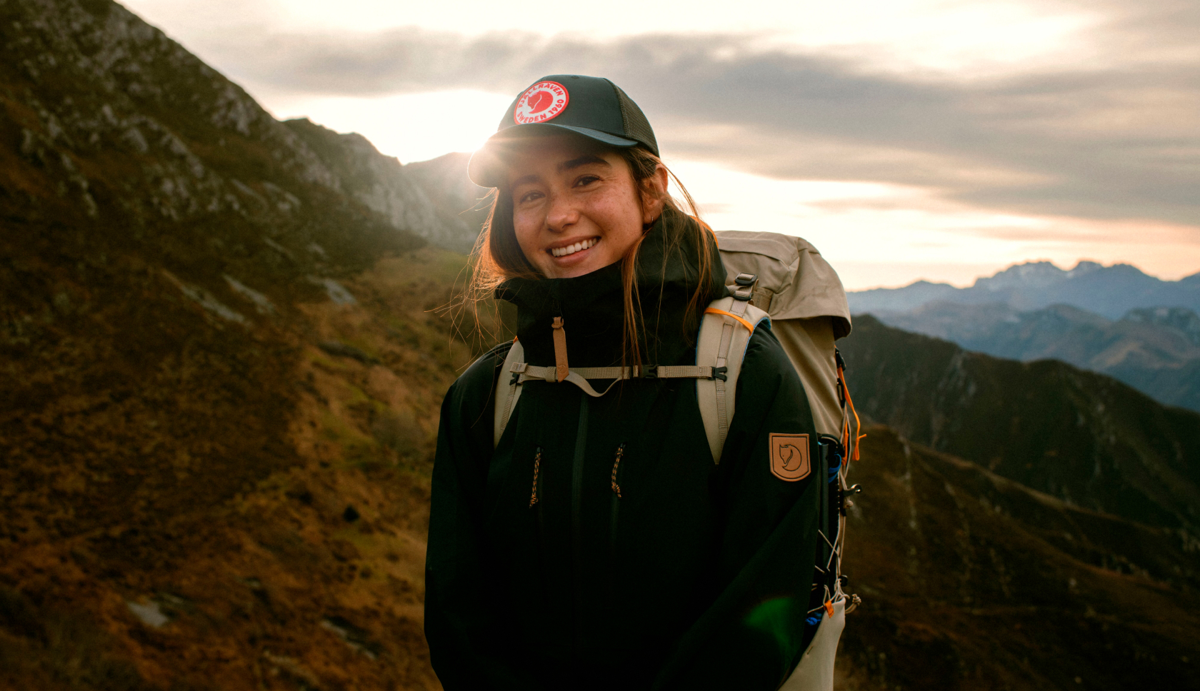 Vrouw in fjallraven jas met backpack om staat in bergachtig gebied en lacht naar de camera