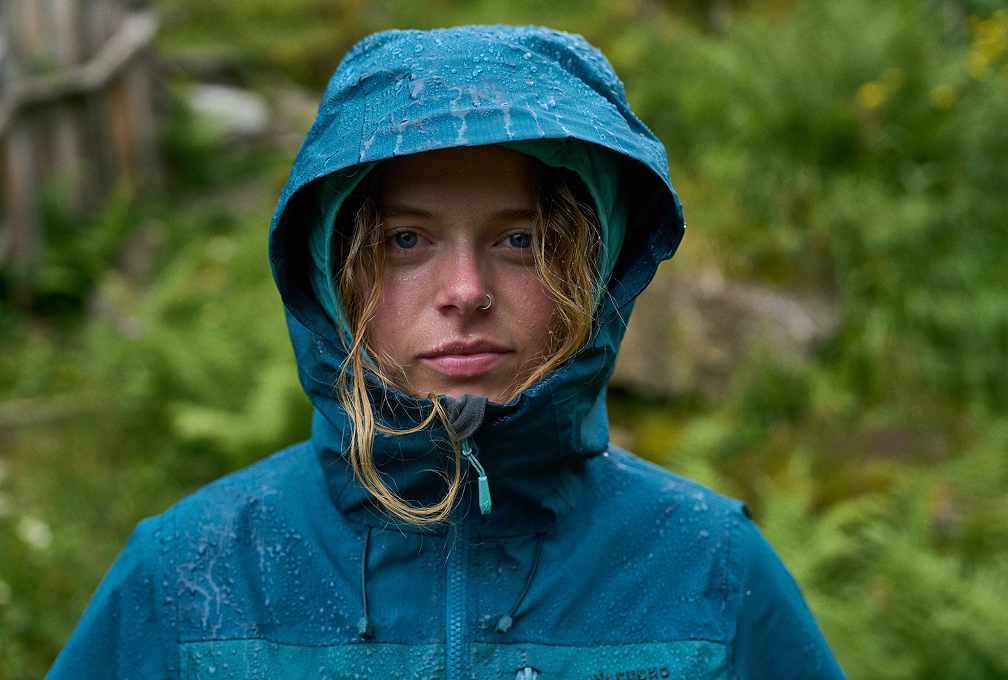vrouw met blauwe regenjas en capuchon op kijkt serieus in de camera