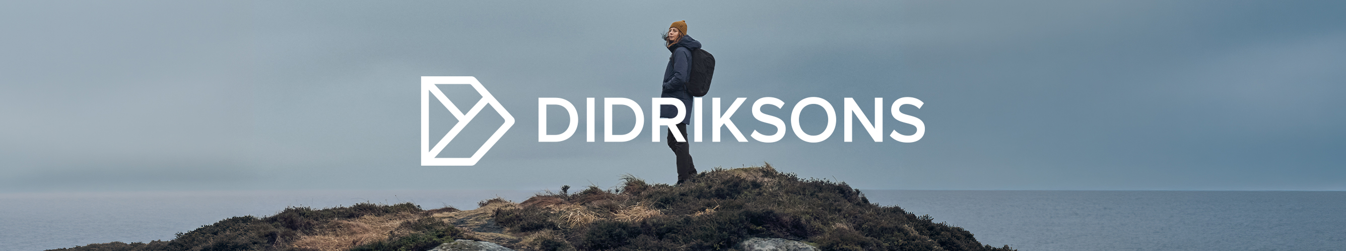 Wandelaar in Didriksons-outdoorkleding staat op een rotsachtige kust, met het Didriksons-logo centraal in beeld.