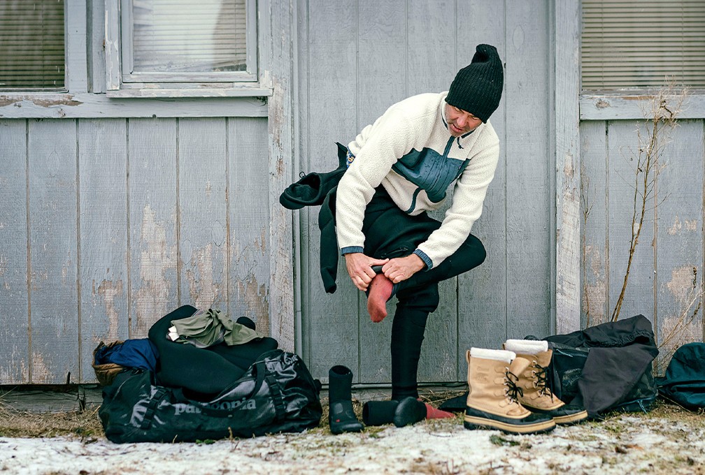 Wandelaar in Patagonia-outdoorkleding trekt buiten bij een houten hut zijn wandelschoenen aan, met reistassen op de grond.